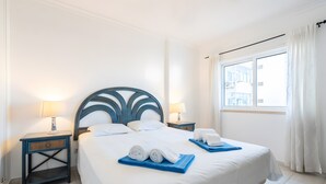 Een net opgemaakt bed met witte lakens, twee nachtkastjes met lampen, en uitzicht buiten door een raam.