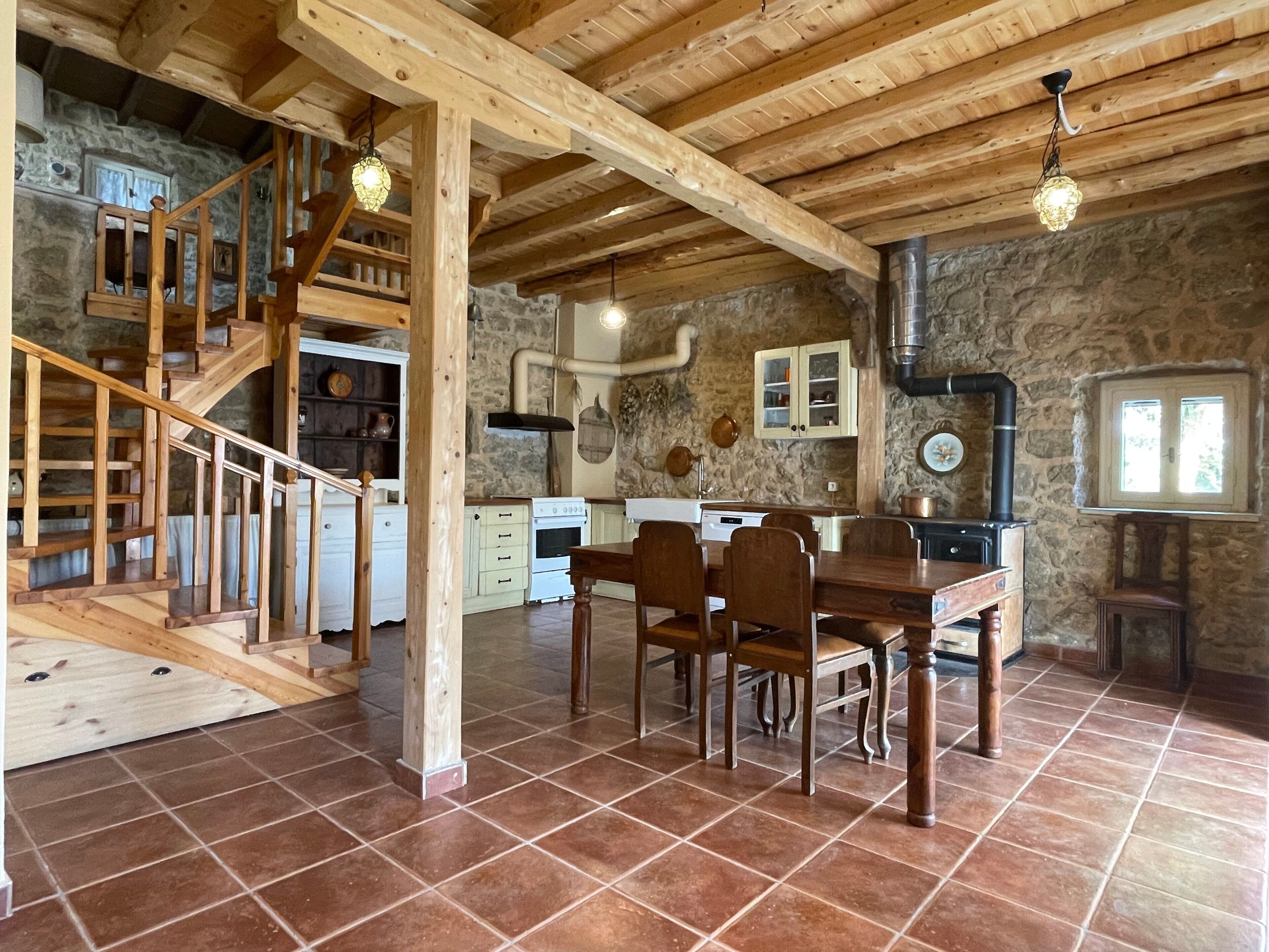 Een rustiek interieur met een houten trap, een eethoek met tafel en stoelen, een keuken met witte kasten en stenen muren.