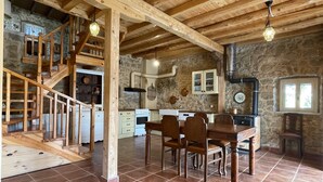 Interior rústico com escadaria de madeira, área de jantar com mesa e cadeiras, cozinha com armários brancos e paredes de pedra.