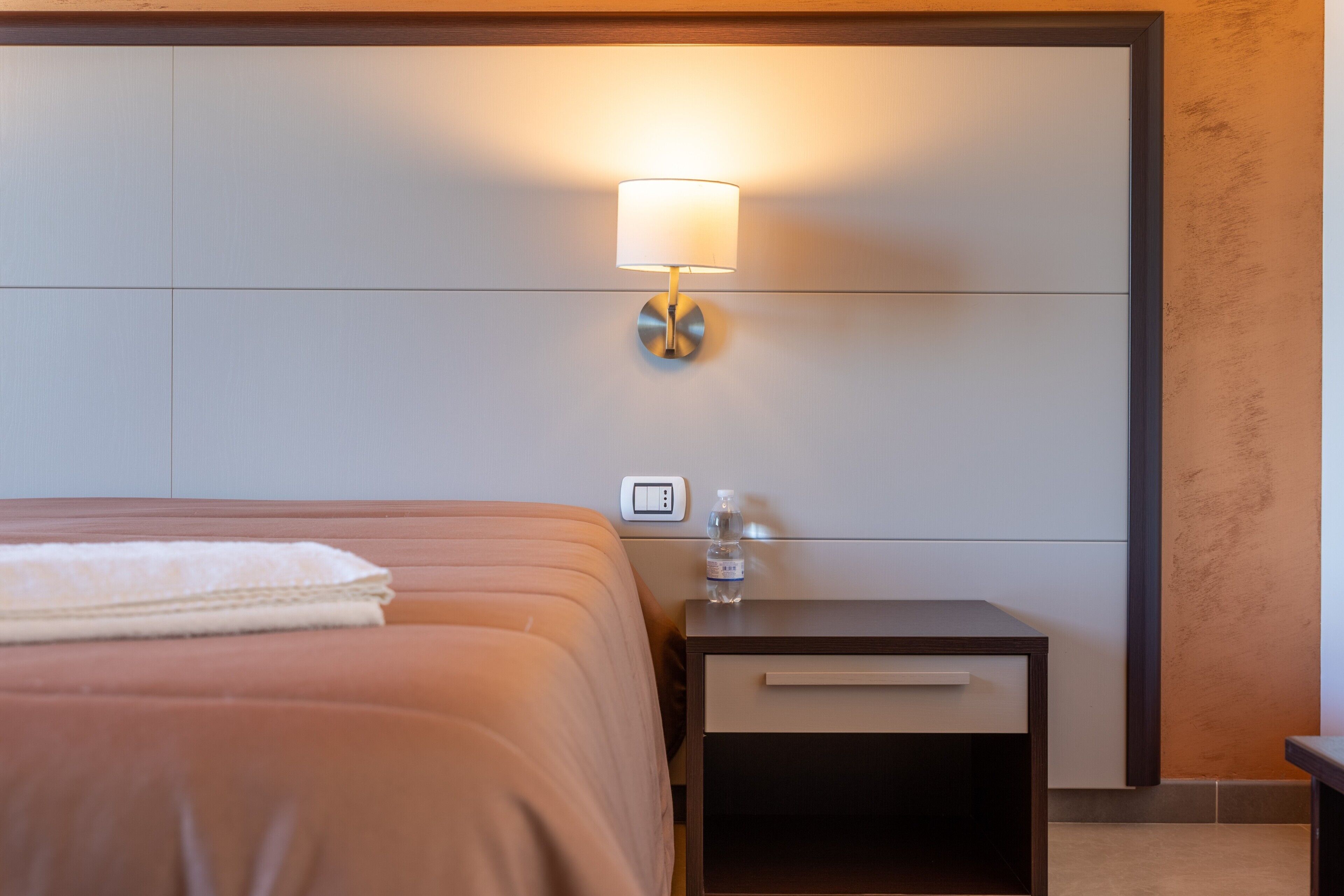 A hotel room with a bed, a nightstand with a lamp, a water bottle, and a thermostat.