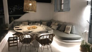 A dining area with a round table, four chairs, a hanging light fixture, a sofa with cushions, and a window with shutters. - Villa 11.7 – Luxus über dem Elia Beach, Mykonos (Elia)