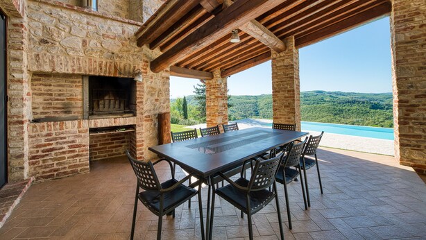 Eine Terrass mit Kamin aus Stein, einer Holzpergola und einem Tisch mit Stühlen.