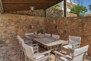 An outdoor patio with a stone wall, a table with chairs, and a hanging light fixture. - Studio Apartment '5' with Shared Garden, Wi-Fi and Air Conditioning (Syvota)