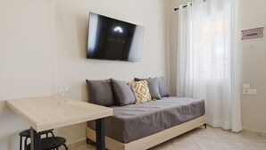 Ein Zimmer mit einem Bett, einem an der Wand befestigten Fernseher, einem Tisch mit zwei Stühlen und einem Vorhangfenster.