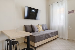 Ein Zimmer mit einem Bett, einem an der Wand befestigten Fernseher, einem Tisch mit zwei Stühlen und einem Vorhangfenster.