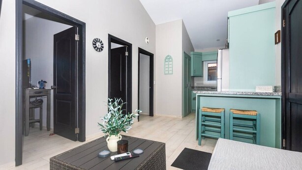 Un espace de vie compact comprenant un canapé, une table basse agrémentée d’une plante, un coin cuisine avec des meubles de couleur turquoise et une petite table à manger avec deux tabourets.