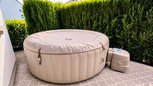 Una bañera de hidromasaje redonda al aire libre con tapa, unidad de control y una unidad más pequeña a su lado, ubicada en un patio con diseño y un seto al fondo.