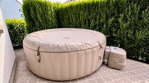 A round outdoor hot tub with a cover, a control unit, and a smaller unit beside it, set on a patterned patio with a hedge in the background.