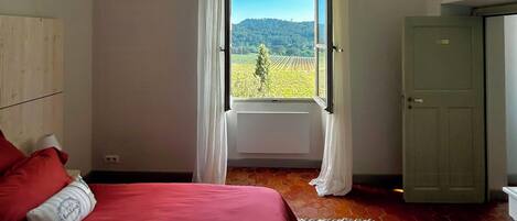 A bedroom with a large bed, a view of a vineyard through an open window, and a door to the right.
