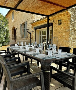 Un espace extérieur aménagé pour les repas, avec des tables et des chaises en osier, un bâtiment en pierre en arrière-plan et une pergola au-dessus.