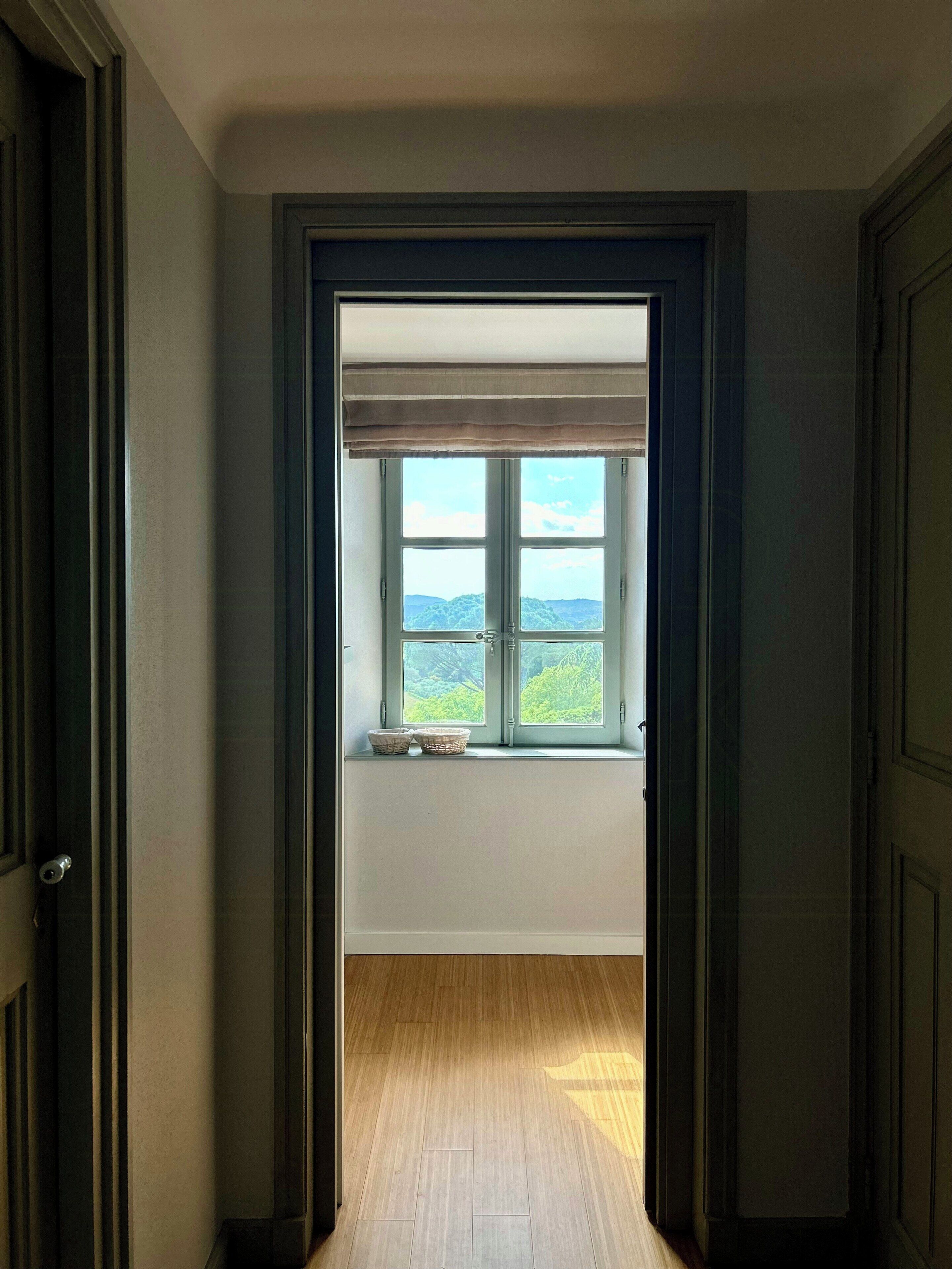 Een gang die uitkomt bij een kamer met een raam, een deur en een houten vloer.