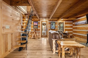Ein Holzblockhaus-Interieur mit einer Holztreppe, einer Couch, einem Esstisch und einem Kamin.