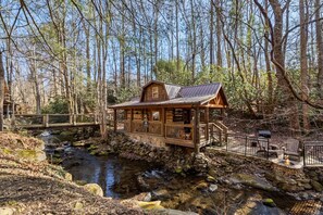 Eine Holzhütte mit Steinunterbau, einer Holzterrasse und einem kleinen Bach.