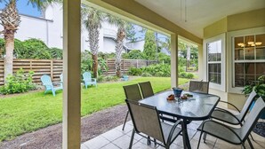 Un patio techado con mesa y sillas, un ventilador de techo y vista a un jardín trasero con palmeras y una cerca de madera.