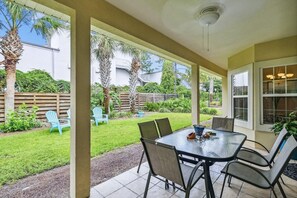 Eine überdachte Terrasse mit Tisch und Stühlen, einem Deckenventilator und Blick auf einen Garten mit Palmen und einem Holzzaun.