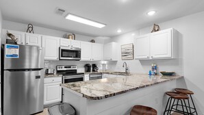 A modern kitchen with white cabinets, stainless steel appliances, a marble countertop, and bar stools. - Grand Caribbean East 112- Crystal Blu (Destin)