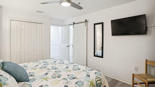 A bedroom with a bed, a ceiling fan, a TV, a mirror, and a door leading to another room. - Grand Caribbean East 112- Crystal Blu (Destin)