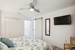 A bedroom with a bed, a ceiling fan, a TV, a mirror, and a door leading to another room. - Grand Caribbean East 112- Crystal Blu (Destin)