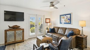A living room with a brown leather sofa, a TV mounted on the wall, a ceiling fan, and a framed picture. - Grand Caribbean East 112- Crystal Blu (Destin)
