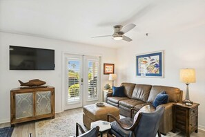A living room with a brown leather sofa, a TV mounted on the wall, a ceiling fan, and a framed picture. - Grand Caribbean East 112- Crystal Blu (Destin)