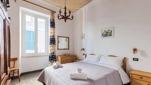 Una camera da letto con un letto, un comodino in legno, una cassettiera, un lampadario, una finestra con tende e un quadro appeso al muro.