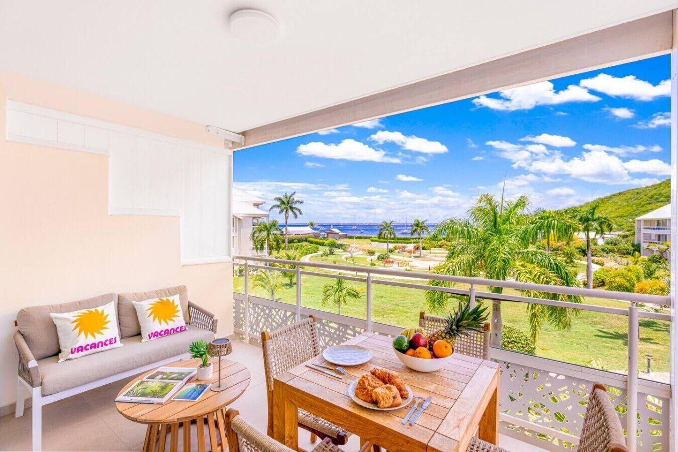 Un balcon donnant sur un paysage tropical, une table à manger avec des assiettes et un saladier rempli de fruits, et un canapé agrémenté de coussins aux motifs solaires.