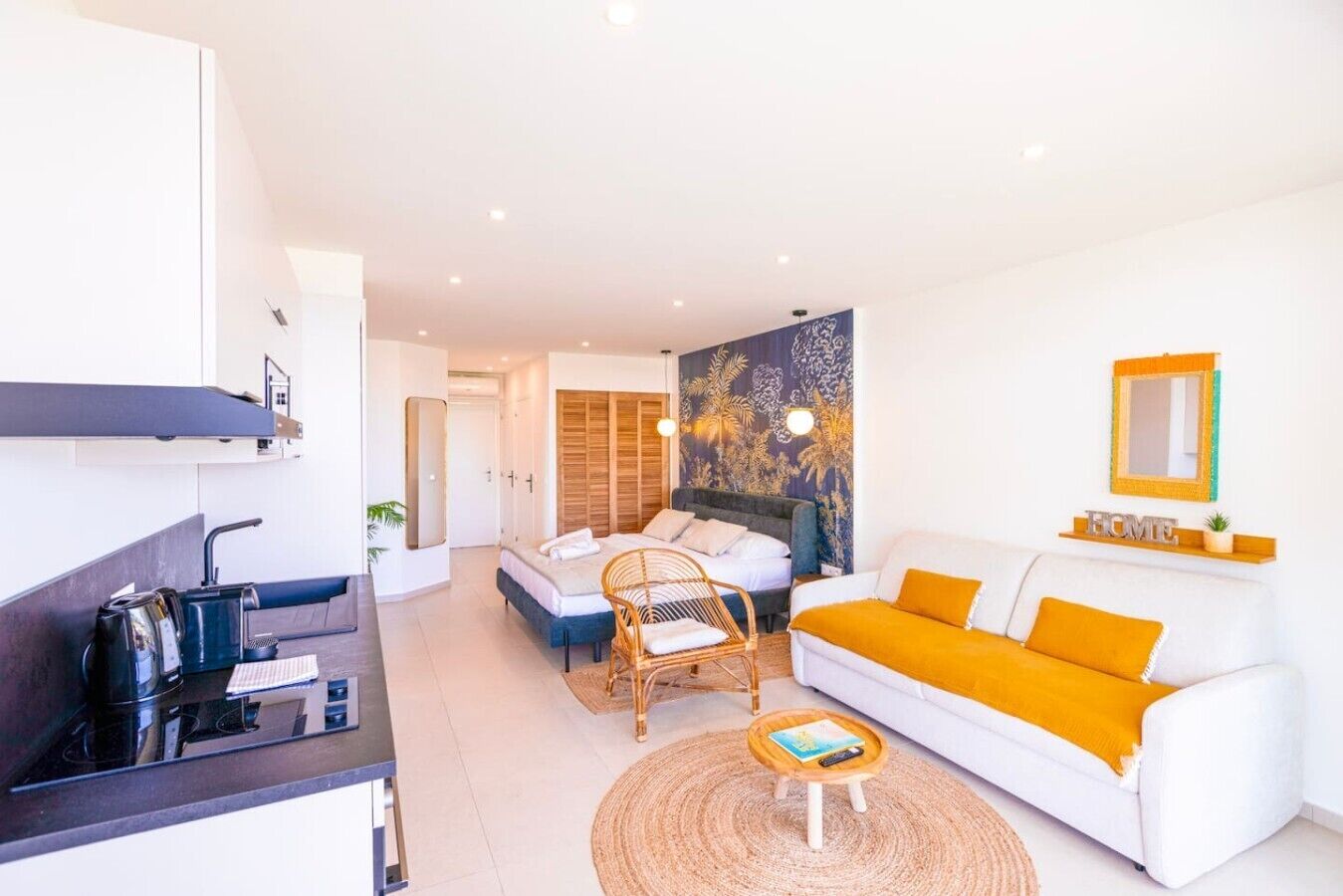 Une chambre d’hôtel moderne dotée d’une kitchenette, d’un lit avec une tête de lit, d’un canapé avec des coussins orange et d’un mur orné d’une tapisserie décorative.