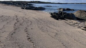 Ein Strand mit felsigem Ufer, blauem Himmel und Hügeln im Hintergrund.