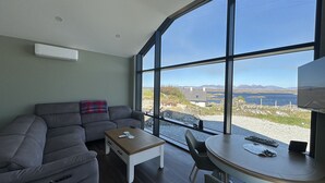 Ein modernes Wohnzimmer mit einer Couch, einem kleinen Tisch und einer Essecke mit einem Stuhl. Große Fenster bieten Blick auf das Meer und ferne Hügel.