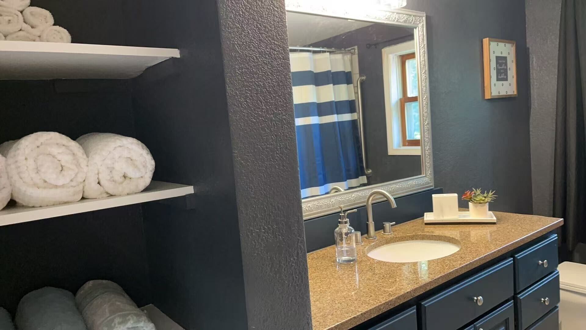 Un bagno con una parete scura, un lavandino bianco, uno specchio e un porta asciugamani con asciugamani arrotolati.
