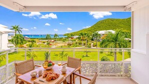 A balcony with a table, chairs, and breakfast items; a view of a beach and greenery; and a white building in the background. - DDL 623, sea view, serenity and comfort (Anse Marcel)