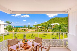 Ein Balkon mit Tisch, Stühlen und Frühstücksgebäck; Blick auf einen Strand und viel Grün; ein weißes Gebäude im Hintergrund.