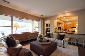 A living room with a sofa, ottoman, and a dining area with a kitchen in the background. - Luxury Power Ranch Estate- Pool, Spa & Golf Green (Gilbert)