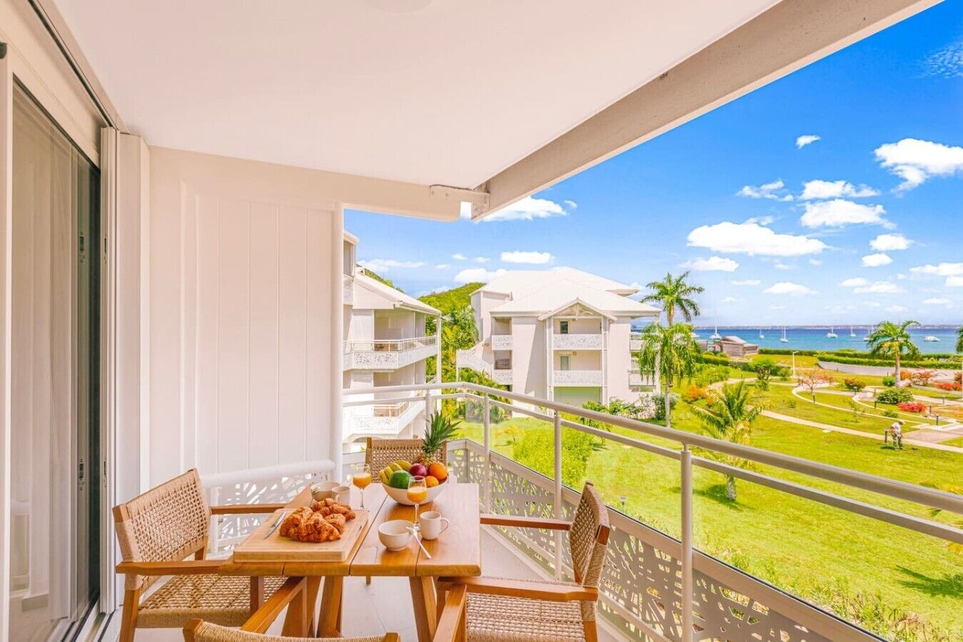 A balcony with a table set for breakfast, overlooking a resort area with buildings and a view of the sea.