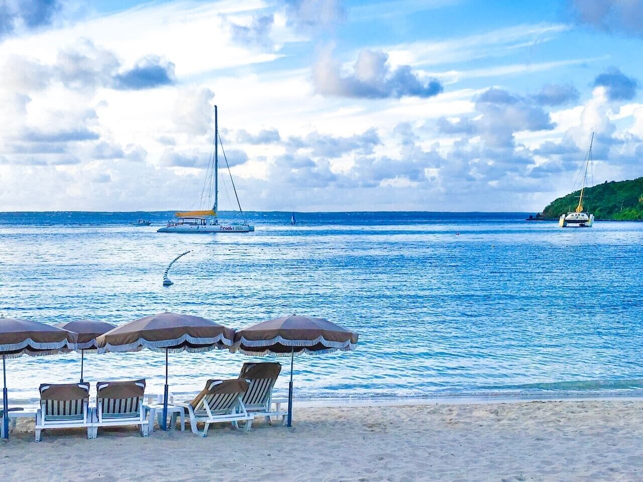 Una spiaggia con sedie a sdraio e ombrelloni, una barca a vela e una piccola isola sullo sfondo, ricca di vegetazione.