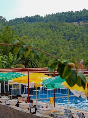 Zona junto a la piscina con sombrillas, sillas de descanso y vista a un paisaje montañoso boscoso.