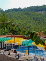 Zona junto a la piscina con sombrillas, sillas de descanso y vista a un paisaje montañoso boscoso.