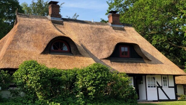 Ein reetgedecktes Häuschen mit zwei Schornsteinen, einem kleinen Fenster mit rotem Rahmen und einer weißen Tür.