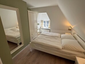 Ein Schlafzimmer mit Bett, Kommode, Spiegel, Fenster mit Vorhängen und einer Lampe.
