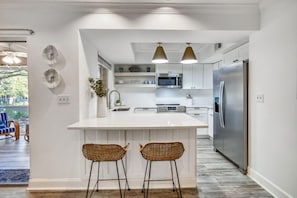 Eine moderne Küche mit weißen Schränken, einer zentralen Kochinsel mit zwei Barhocker und einem Edelstahlkühlschrank, beleuchtet durch Pendelleuchten.