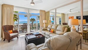 A spacious living room with a sofa, armchair, and coffee table, large windows with a view of the ocean, and a ceiling fan. - Jade East 130- Jade Gem (Destin)