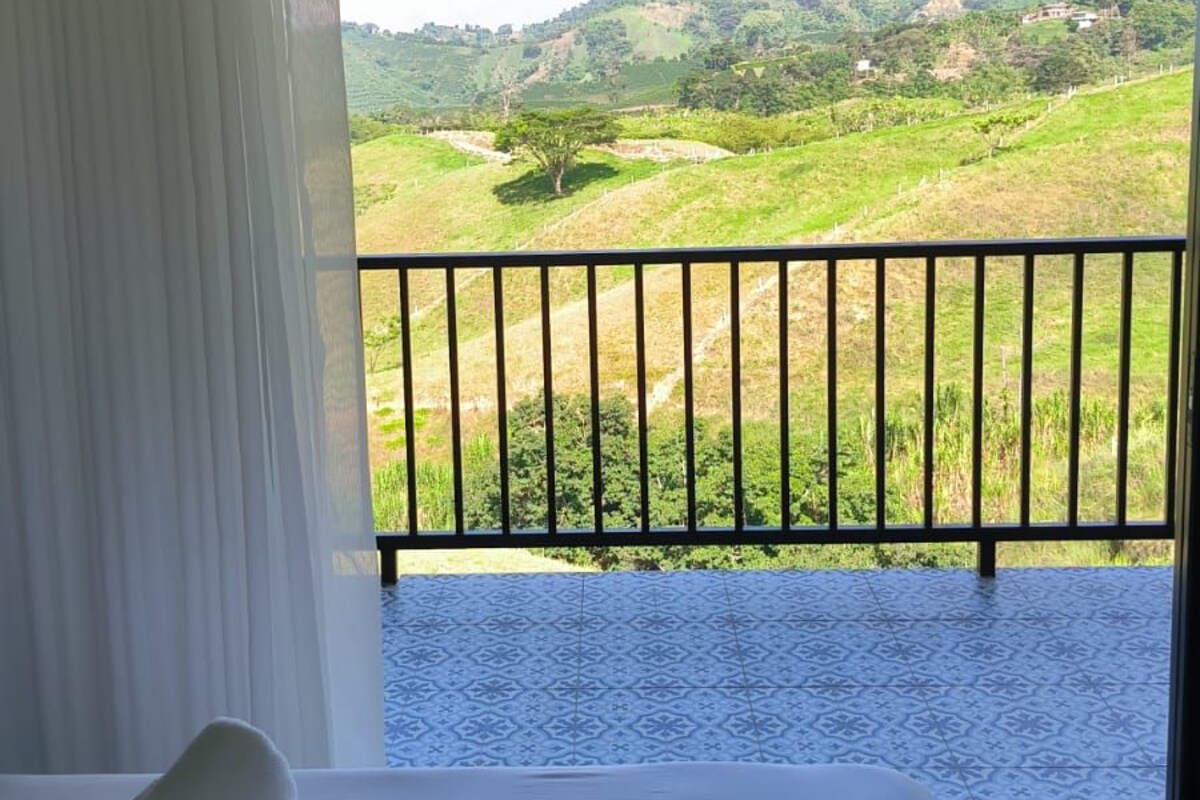 A hotel room with a bed, a window with a view of a green landscape, and a balcony with a railing.