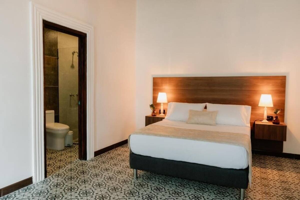 A hotel room with a bed, two bedside lamps, a wooden headboard, a patterned floor, and a bathroom visible through an open door.