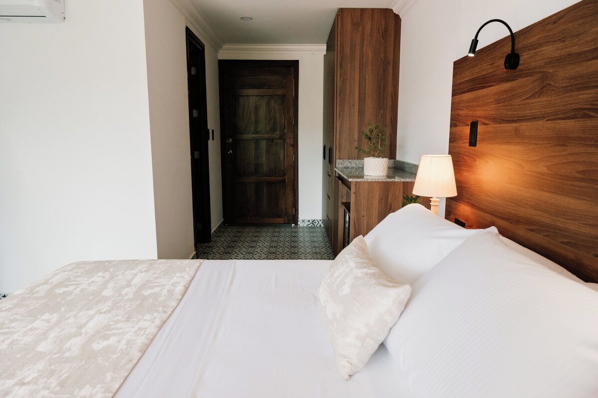 A hotel room with a bed, a nightstand with a lamp, a wooden headboard, and a door leading to another room.