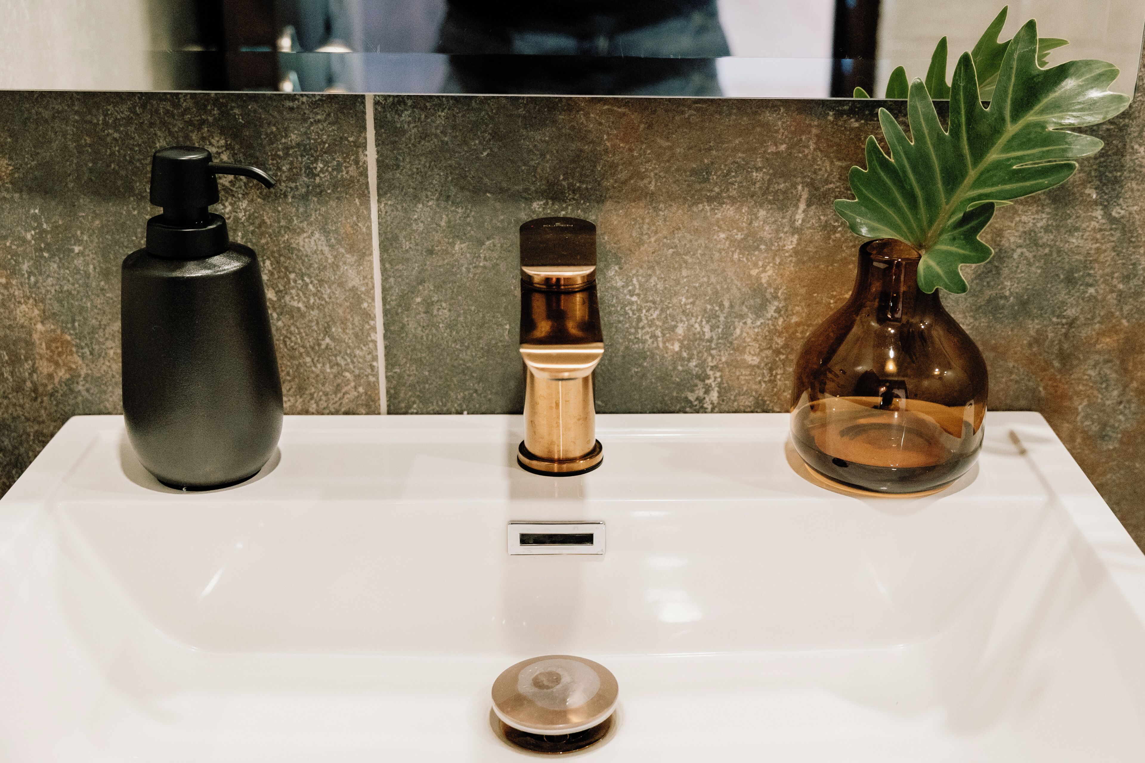 Un lavabo de salle de bain avec un distributeur de savon noir, un robinet doré et un vase transparent contenant une plante.