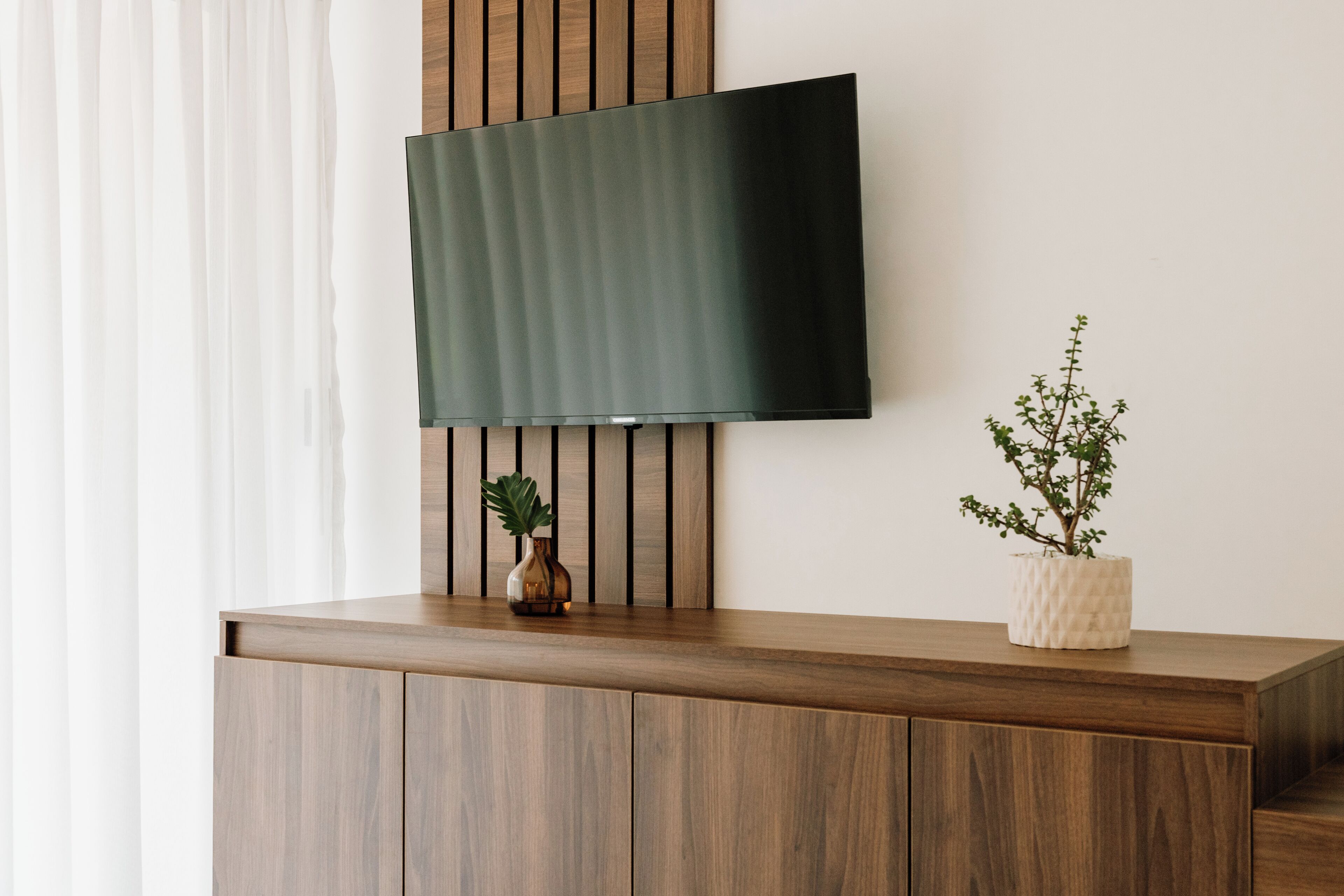 A flat-screen TV mounted on a wooden wall with a plant in a vase on a wooden counter below.