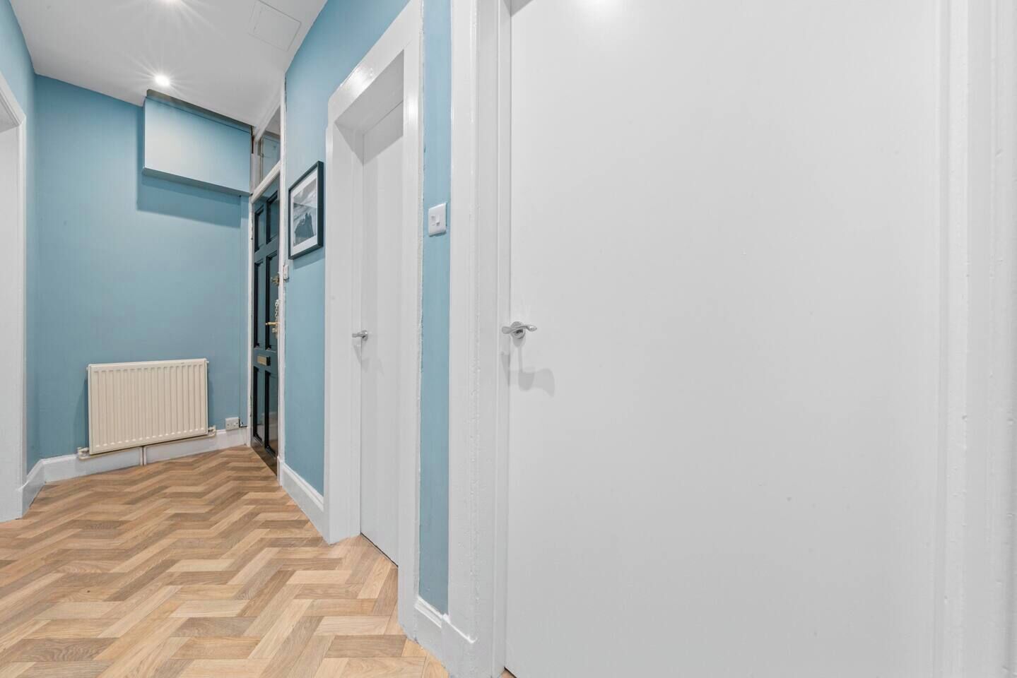 A hallway with a white door, a radiator, and a herringbone-patterned floor.