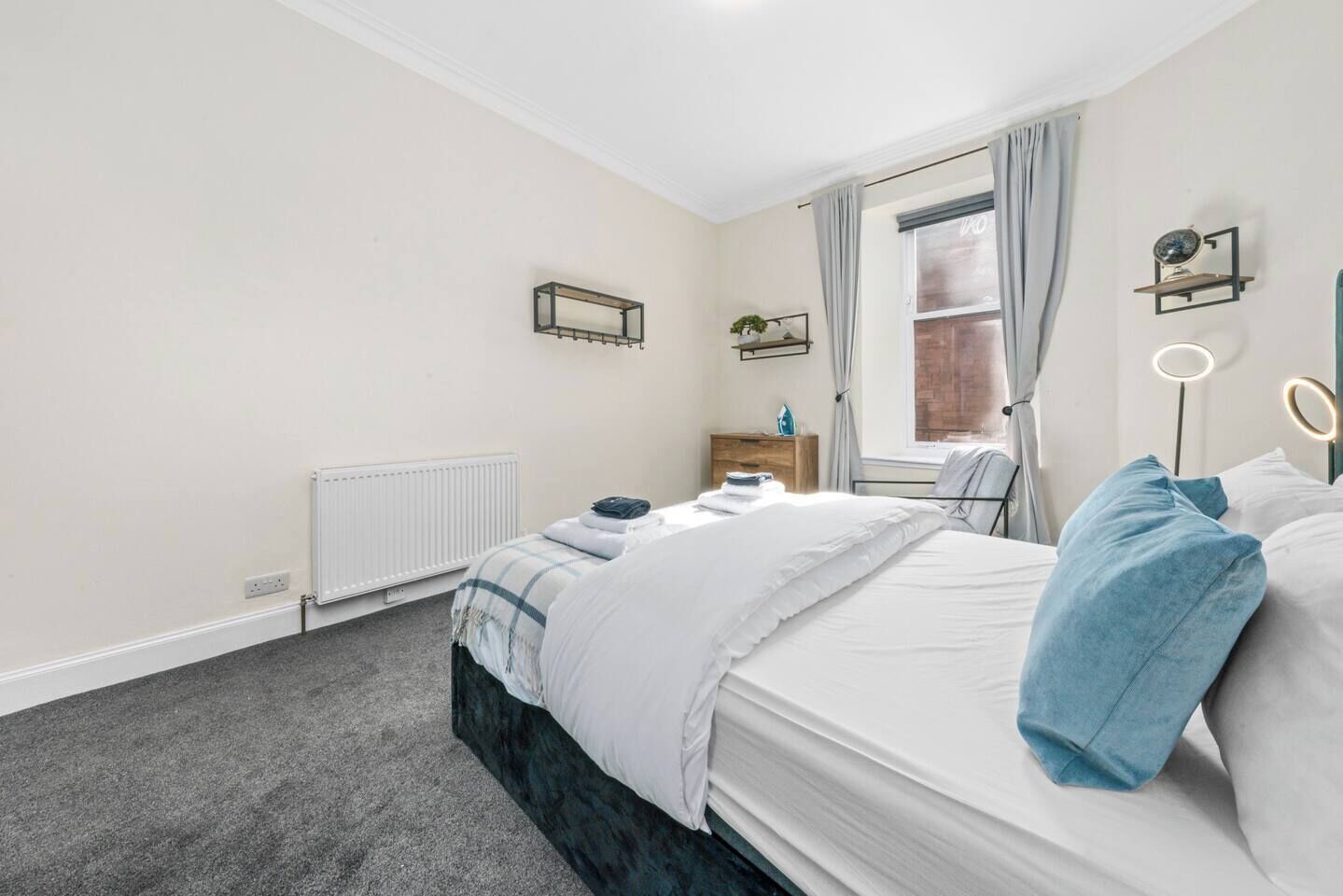 A bedroom with a bed, a radiator, a window with curtains, a bedside table, and a wall-mounted shelf.