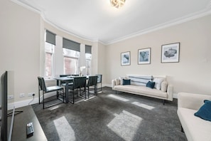 A living room with a sofa, a dining table with chairs, a TV, and artwork on the wall.