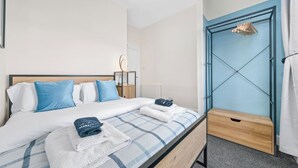Ein ordentlich bezogenes Bett mit einer blau-weiß karierten Tagesdecke, einem hölzernen Kopfteil und einem Nachttisch mit Lampe und Spiegel.